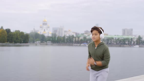 Young Asian Boy in Headphones Running along Urban Riverside