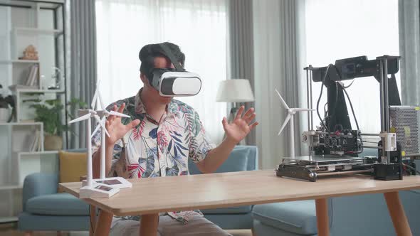 Asian Man Wearing VR Glasses While Experimenting With Wind Turbine Model And 3D Printer At Home