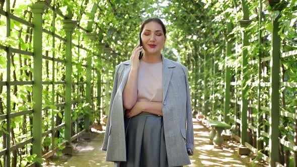 Young Woman Talking on the Phone in an Arched Corridor of Trees in the Garden