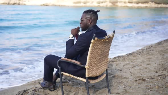 Wide Shot of Handsome Elegant African American Man Sitting on Wicker Chair on Sandy Mediterranean