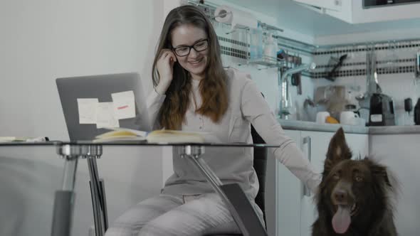 Young Woman At Home Sitting At The Table Using Her Laptop Close To Her Dog.