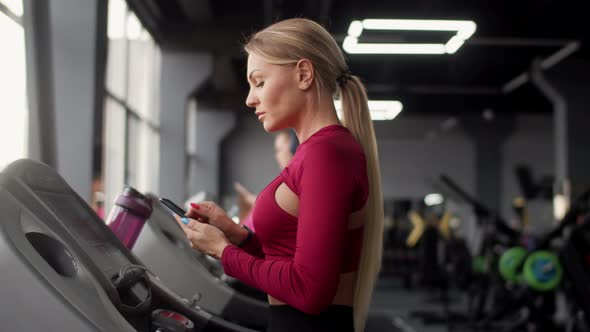 Athletic Woman Does Cardio Training on Treadmill in Gym and Looks at Smartphone Side View