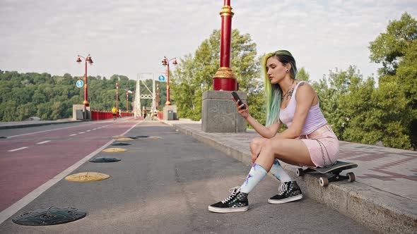 Female Teen Skateboarder with a Bright Appearance Tattoo and Colored Hair Sits in a City Park Near