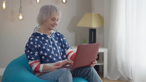 Modern Attractive Mature Slim Woman Listening to Music Sitting with Laptop on Bag Chair Indoors