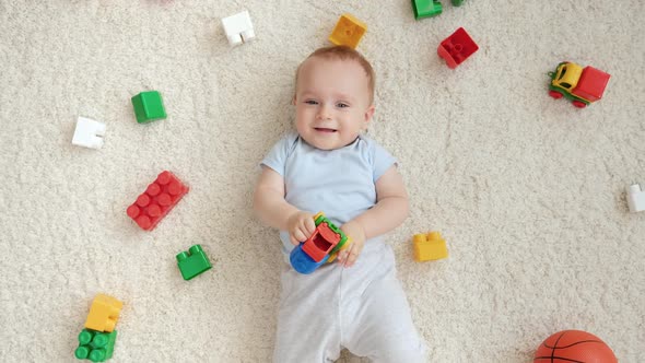 Happy Smiling Baby Boy with Lots of Toys Lying on Carpet and Looking Up in Camera
