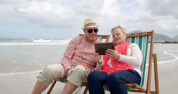 Front view of old caucasian senior couple using digital tablet and sitting on sunlounger a beach 4k