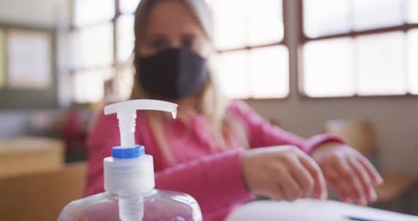 Girl wearing face mask sanitizing her hands while sitting on her desk at school