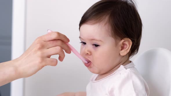 Baby Girl Eating Blend Mashed Food Sitting on High Chair Mother Feeding Child Hand with Spoon for