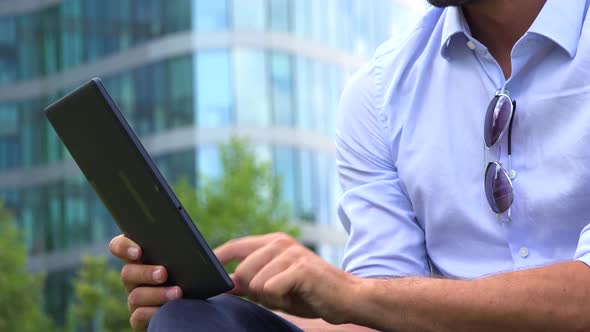 A Young Handsome Businessman Works on a Tablet - Closeup - an Office Building in the Background