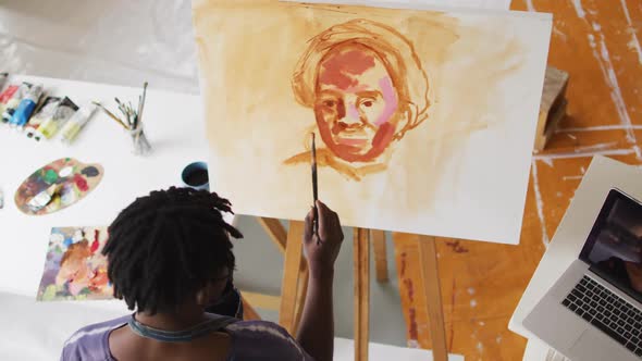 African american male artist wearing face mask painting on canvas looking in laptop at art studio