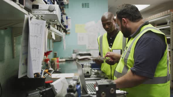 A Caucasian and an African American male factory workers standing, in discussion, using a computer 