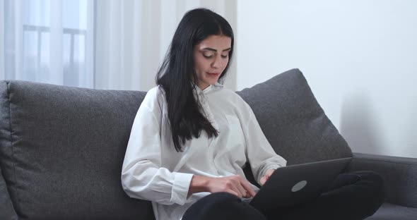 Young Woman Freelancer Is Sitting on the Couch and Working on a Laptop Types Text on the Keyboard