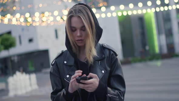 Young Girl Uses a Smartphone Near a Modern Residential Area Evening in the City Blurry Lights on