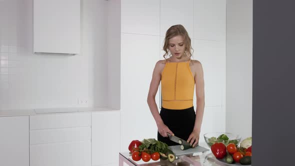 Young Woman Slicing Cucumber For Vegetable Salad. White Modern Kitchen.