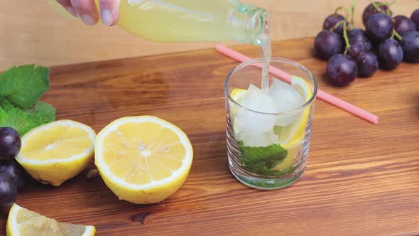 Pouring lemonade from bottle into glass with mint, ice cubes and lemon slices, close up view