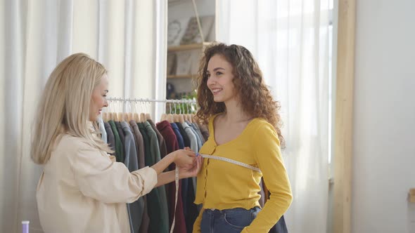 Young Blonde Tailor Measuring Woman Back to Make Custom Made Suit for Pretty Young Client in Her