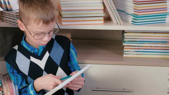 Seven-year-old Boy with Glasses Writes Something in a Sketchbook Sitting Among the Books