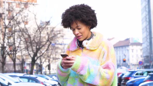 Young beautiful curly hair black woman outdoor in the city smiling using smartphone