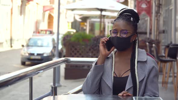 Young Black Woman in Mask Talks on Phone in Street Cafe. Beautiful Female Using Mobile Phone To Talk