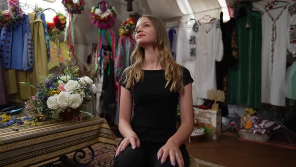 Curios Young Woman Sitting in Shop with Ukrainian Embroidered Dresses Looking Around