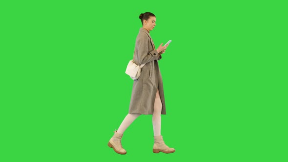 Young Woman in Coat Walks Scrolling and Typing Something on Smartphone on a Green Screen Chroma Key