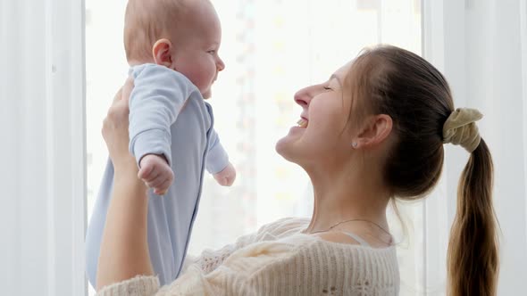 Portrait of Young Mother Lifting Up and Smiling at Her Little Baby Son in Front of Big Window at