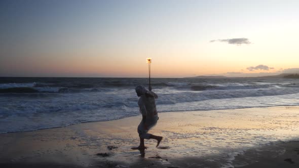 A man fire dancer in silhouette spinning his flaming staff on the beach at sunset with flames and oc