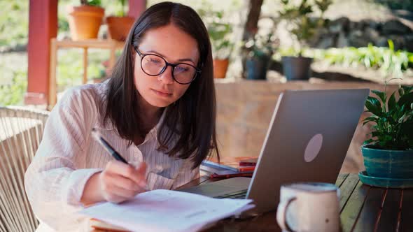 A Young Woman Freelancer Makes Notes in a Notebook and a Laptop