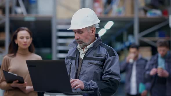 Busy Senior Man in Hard Hat Using Laptop As Cheerful Young Beautiful Woman Approaching Talking and
