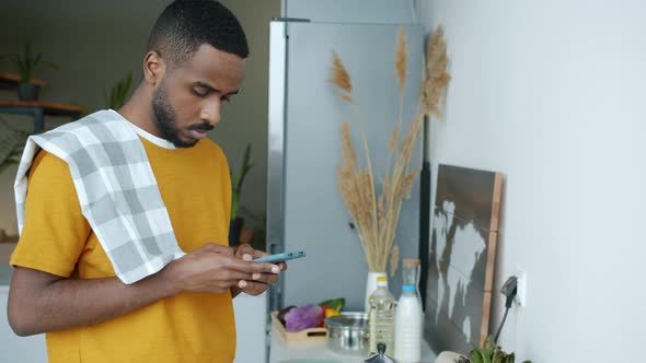 Young Man Using Smartphone and Cooking Breakfast in Kitchen in Modern Apartment