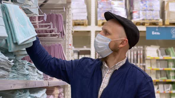 A Masked Man in a Shop with a Shopping Cart Walks Near the Shelves with Goods and Chooses a Blue