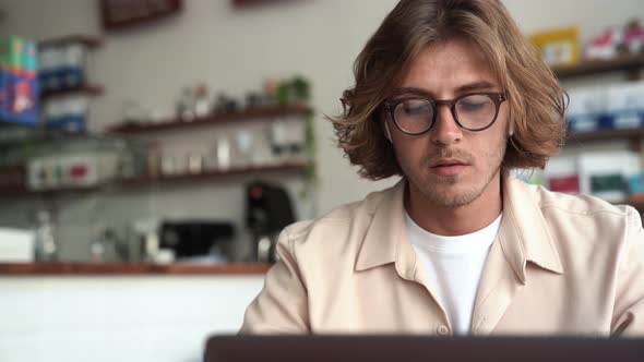 Pensive blond man in eyeglasses working on laptop and looking to the side