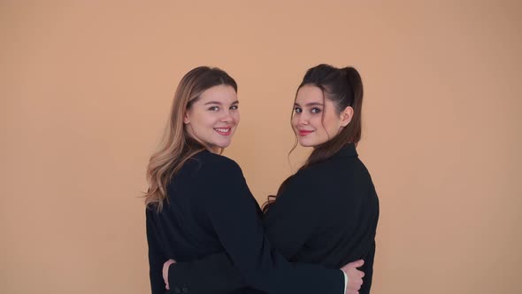 Two Young Business Women Stand in the Studio Backs to Camera Hugging Turns Around and Blows Kiss