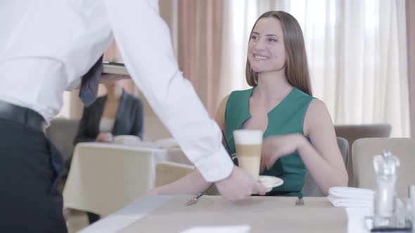 Waiter Bringing Latte for Smiling Brunette Woman in Cafe and To Blond Businesslady at the Background