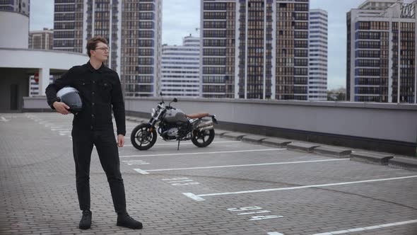 Smart Guy Stands in Front of Motorbike with Helmet at Top of Multilevel Parking