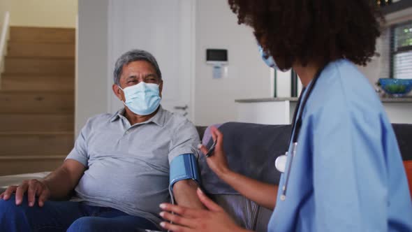 Mixed race female doctor wearing mask and senior man taking blood pressure at home