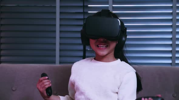 Asian little girl having fun playing video game with virtual reality glasses on sofa at home.
