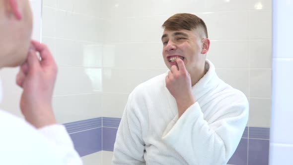 A Young Man in a White Coat Looks in the Mirror at His Teeth. View Through the Mirror