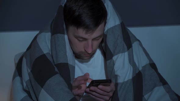 Young Man Cell Phone Addict Wakes Up Late at Night in Bed Wrapped Upside Down in Blanket Using