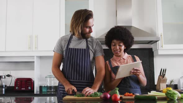 Mixed race couple wearing aprons using digital tablet and chopping vegetables together in the kitche