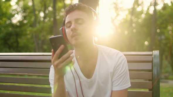 A Young Handsome Guy in a White Tshirt and Headphones Sits on a Bench in the Park at Sunset and