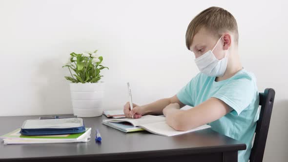 A Young Boy in a Face Mask Does Homework for School at a Table at Home