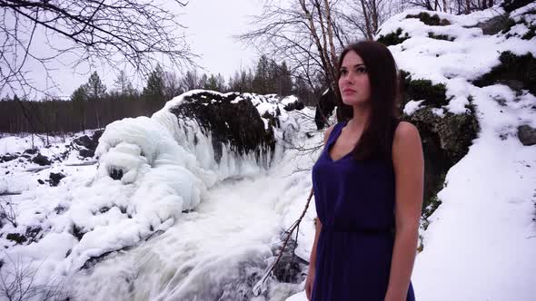 A beautiful girl in a blue dress at a frozen waterfall looks into the distance 4K