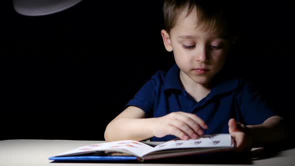 A Child Opens and Reads a Book at Home Sitting at Night at a Table That Is Lit By a Lamp