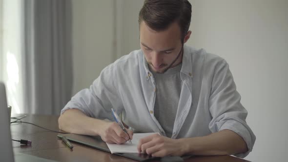 Man Sitting at the Table Writing Something on the Paper at Home