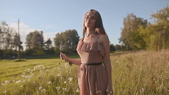 Beautiful Girl with Long Hair Goes in a Green Meadow and Touch the Flowers