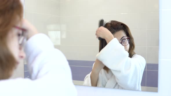 Young Woman with Glasses Combing Her Hair in Front of a Mirror. A Girl in a White Bathrobe in Front