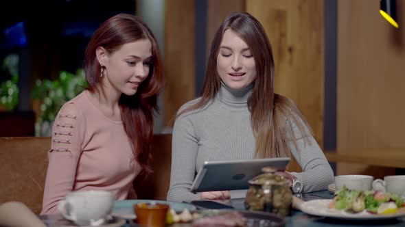 Middle Shot of Two Beautiful Confident Young Women Surfing Internet on Tablet and Talking in Cafe