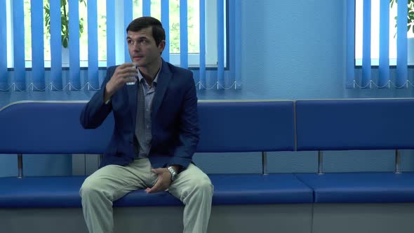 The Adult Man Sitting in the Corridor in Modern Private Clinic Drinking Water From the Glass Waiting