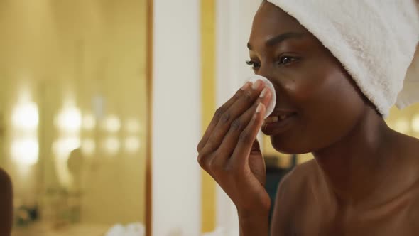 Smiling african american woman with towel using pad on her face and watching in mirror in bathroom